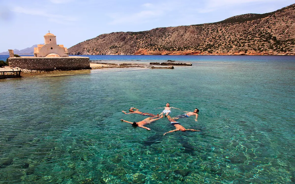 Two people floating in shallow clear water beside a stone building on the shore of a small Aegean island