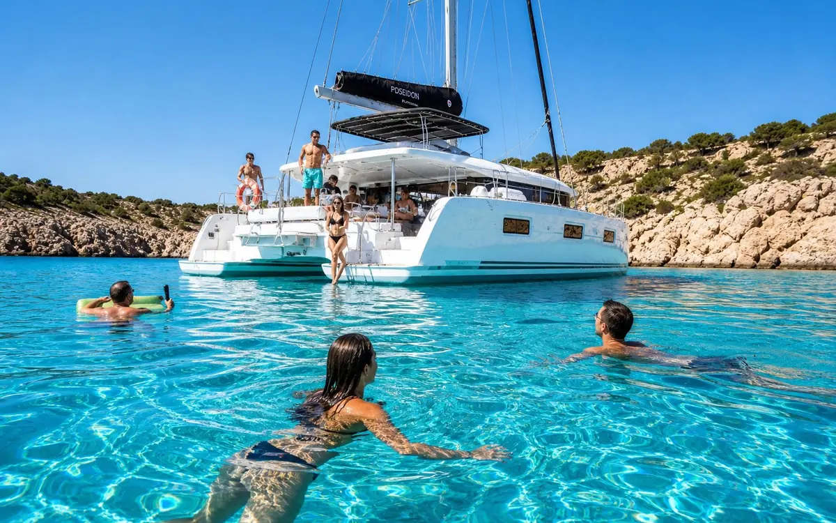 A white catamaran anchored close to a rocky shore in Rhodes with people swimming and floating in the clear water around it