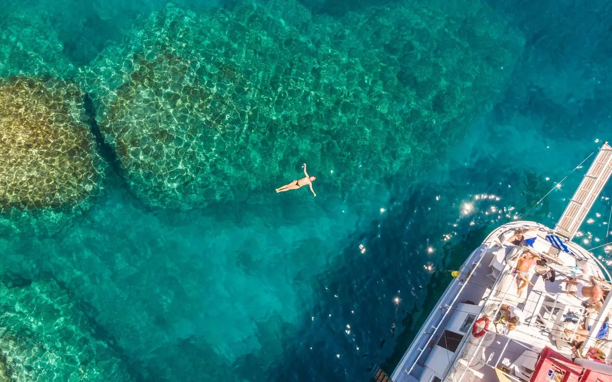 A wooden excursion boat seen from above on clear turquoise water off Rhodes with one swimmer in the water nearby