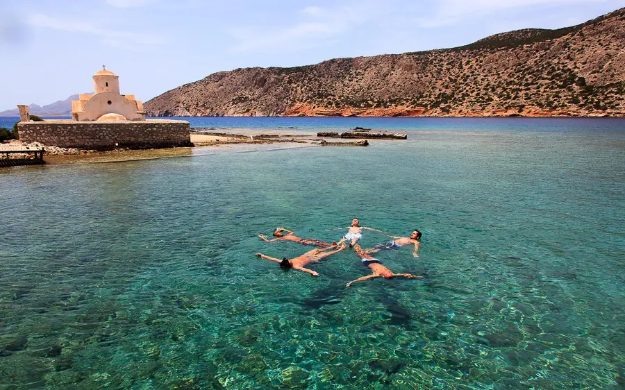 Two people floating on their backs in shallow clear water beside a bare headland with a small stone chapel