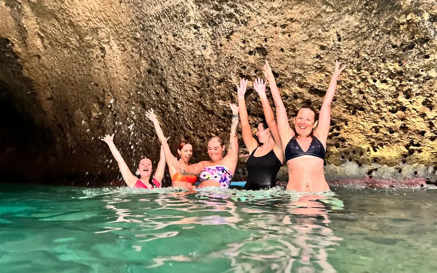 Five people standing chest-deep in clear green water below a rocky cliff, arms raised above their heads