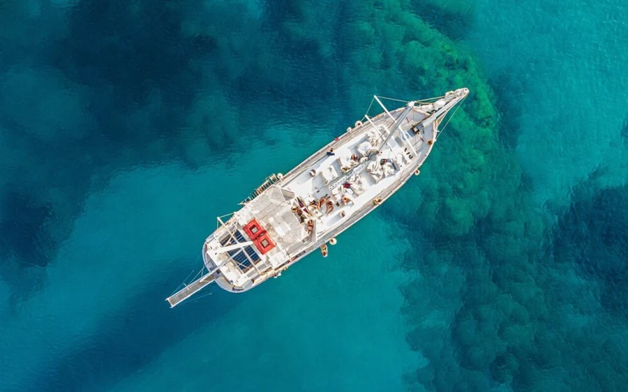 An aerial view of a white wooden boat on turquoise water, its shadow lying on the seabed below