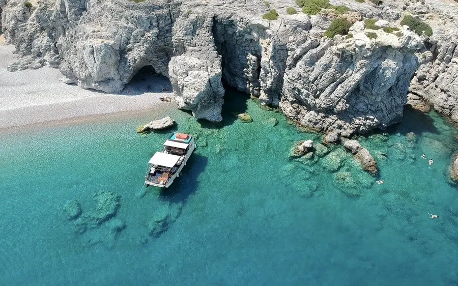 An aerial view of a small white boat anchored beside pale rock cliffs and a narrow pebble beach