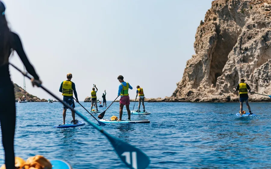 A group on stand-up paddleboards in life jackets paddling on blue water below a tall rock headland