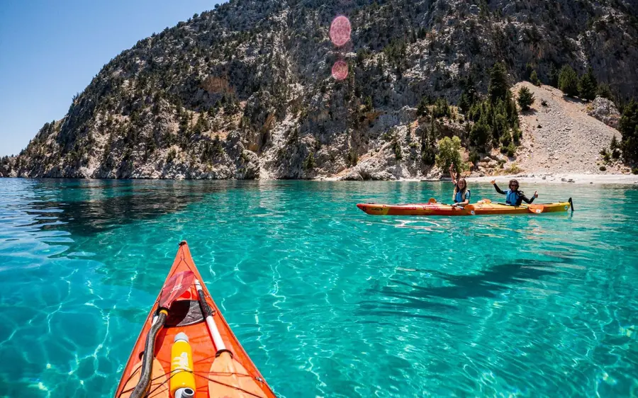 The view forward from an orange sea kayak towards a green cliff and a beach across turquoise water